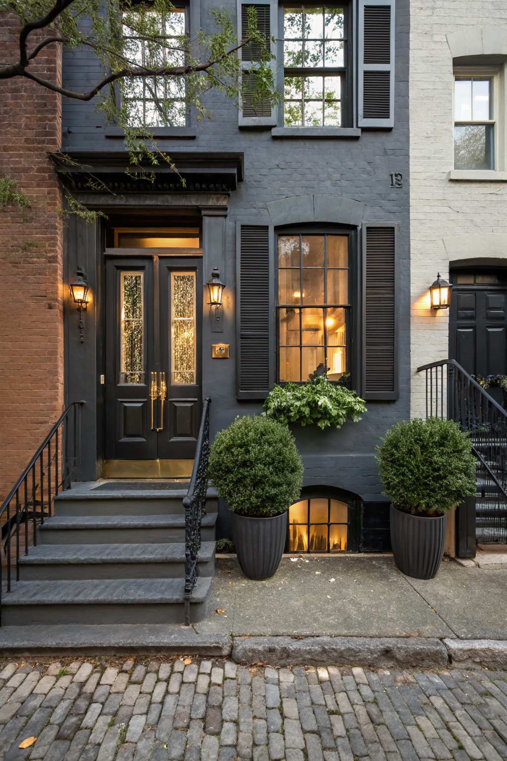 Dark gray painted brick townhouse exterior featuring black double doors with brass hardware and glass panels, flanked by lantern lights, potted boxwood shrubs on steps, and basement window, adjacent to white house on cobblestone street.