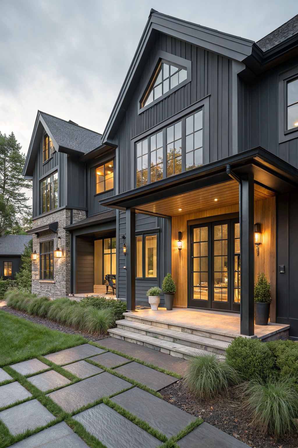 Two-story dark gray vertical board-and-batten house exterior with a covered front porch featuring light wood beams and ceiling, black-framed glass doors and windows, stone entry steps, and low landscaping.