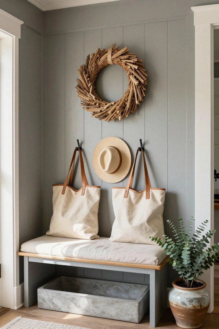 Light gray shiplap wall in an entryway corner with a large circular wreath of twisted driftwood sticks centered above black hooks holding a wide-brim straw hat between two beige canvas tote bags with leather handles, beside a wooden bench with white cushion, metal basket below, and potted eucalyptus plant in terracotta pot.
