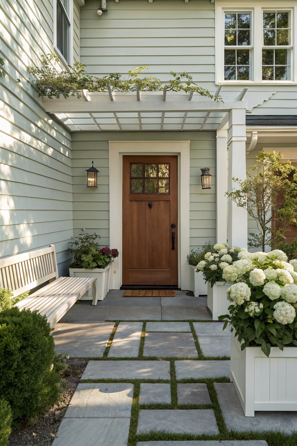 Light green clapboard house exterior featuring a dark brown wooden entry door with glass panels, flanked by white planters filled with white hydrangeas, a wooden bench, lanterns, and a pergola with vines overhead on a stone paver pathway.