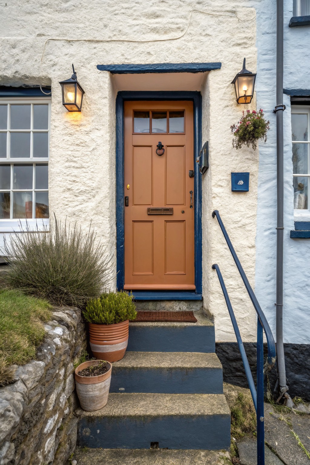 Whitewashed stone house facade with a tan paneled wooden front door in a blue frame, flanked by black lanterns, potted plants, and blue handrail on stone steps.