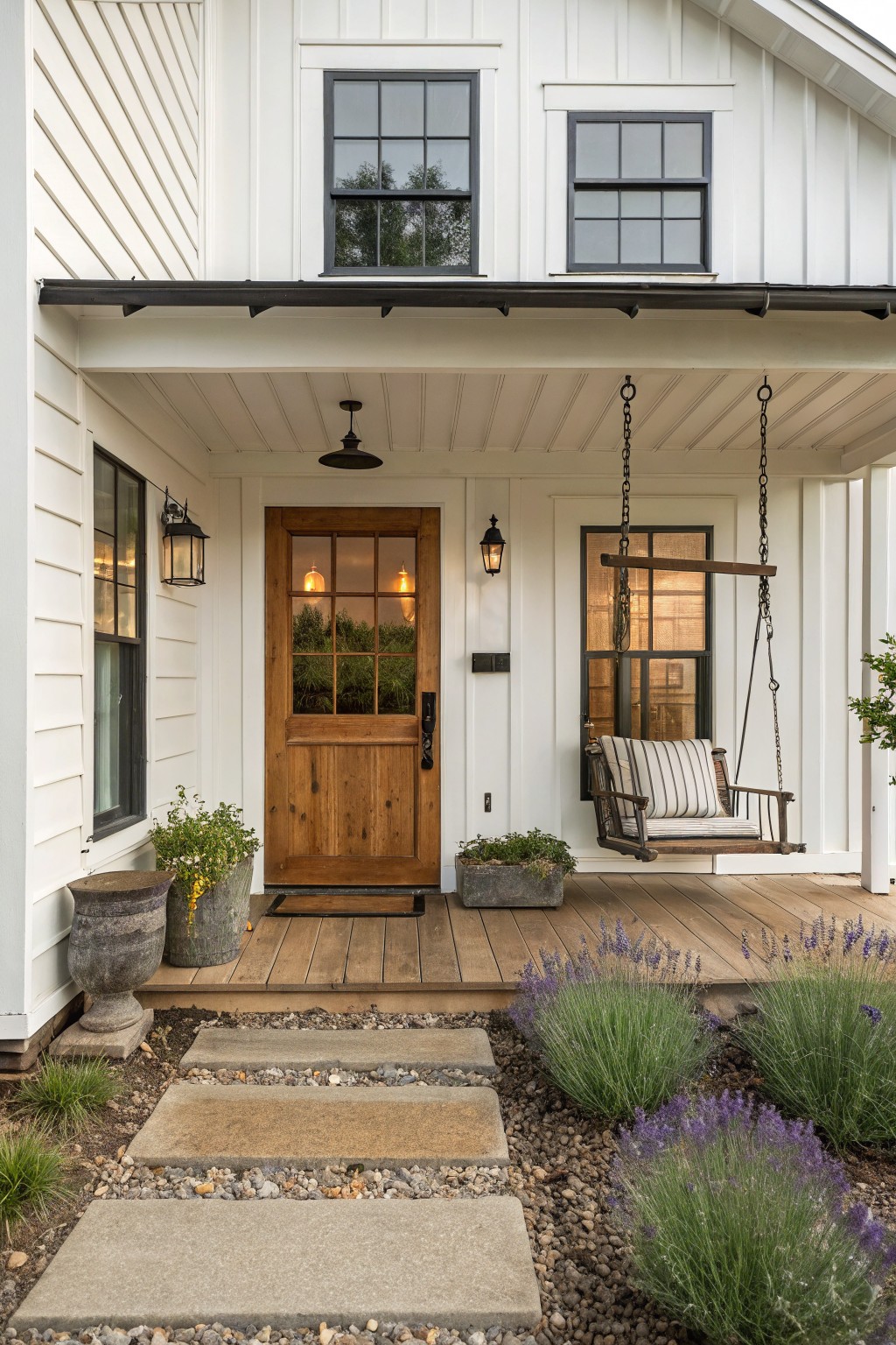 Front exterior of a white board-and-batten house featuring a brown wooden door with glass panels, flanked by lanterns, a porch swing, potted plants, and a stone path edged with lavender bushes.