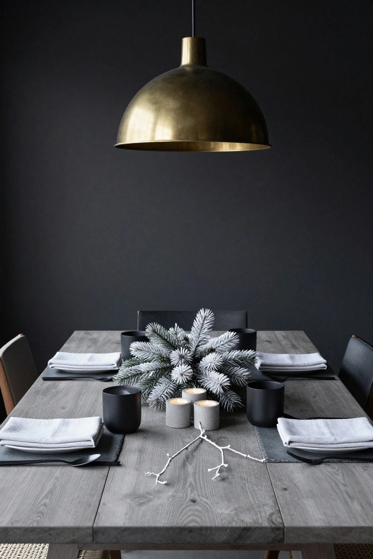 Dark-walled dining room with a rectangular grey wood table set for four, featuring black ceramic mugs, white linen napkins, frosted spruce branches centerpiece, concrete candles, and black chairs, lit by a large brass dome pendant light.