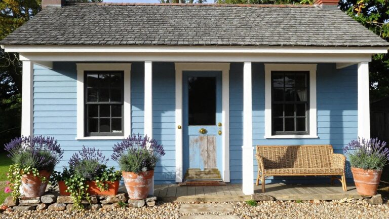 Light blue shingle-style cottage house with white trim and porch, wooden front door, two large terracotta pots of lavender on the wooden porch, and stone wall with gravel landscaping at the base.