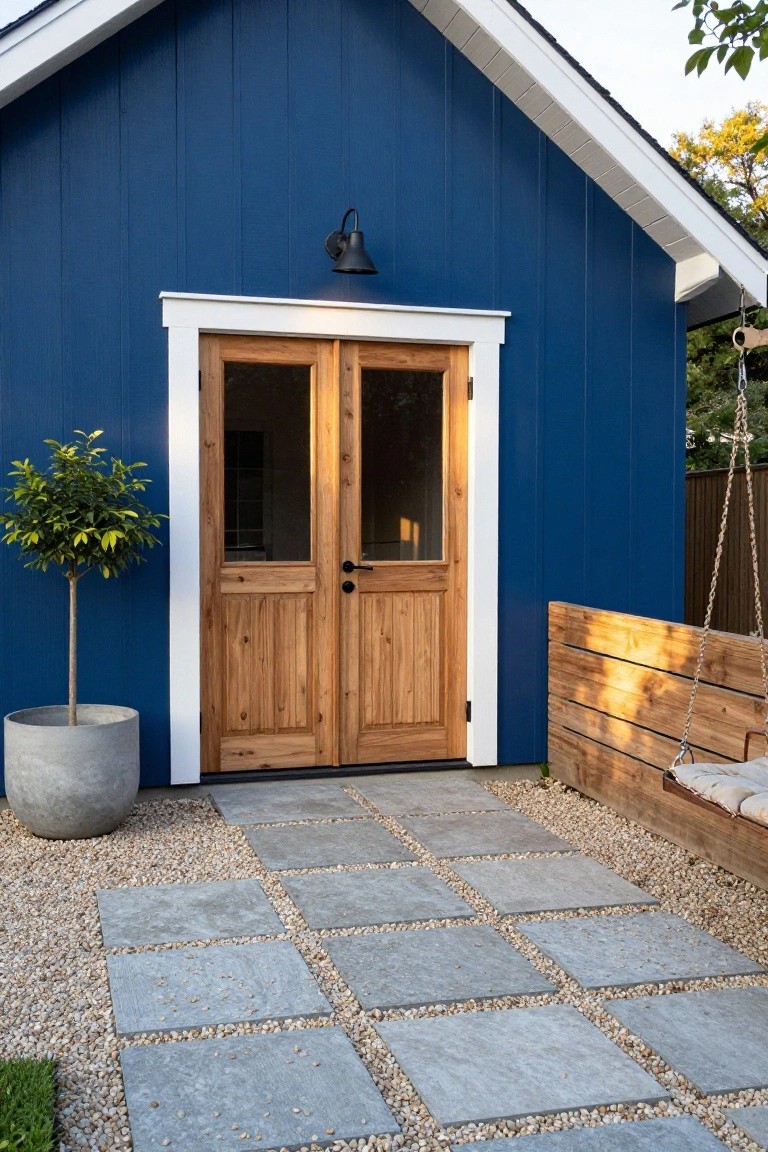 Navy blue shed with white trim and double wooden doors, a potted citrus tree beside it, concrete paver pathway with gravel edges, and wooden swing bench to the right.
