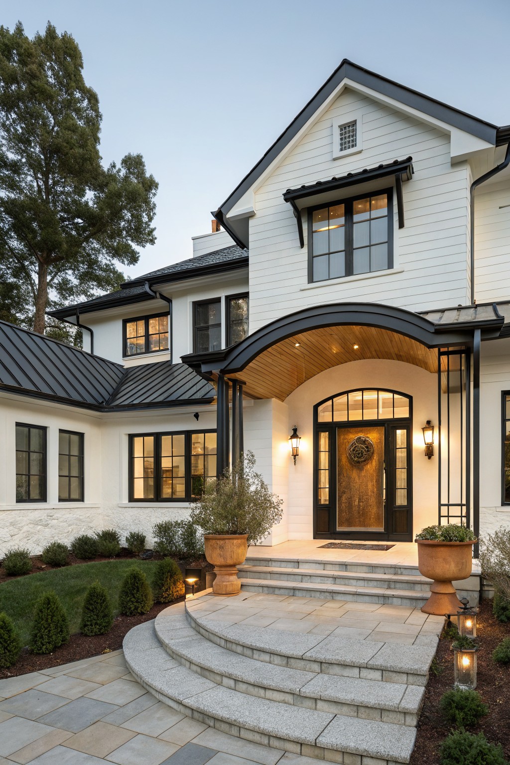 White shiplap house exterior with black metal roof and gable, featuring an arched portico entry with wooden ceiling and black metal supports, wooden door with wreath, curved granite steps, potted plants, boxwoods, and landscape lighting.