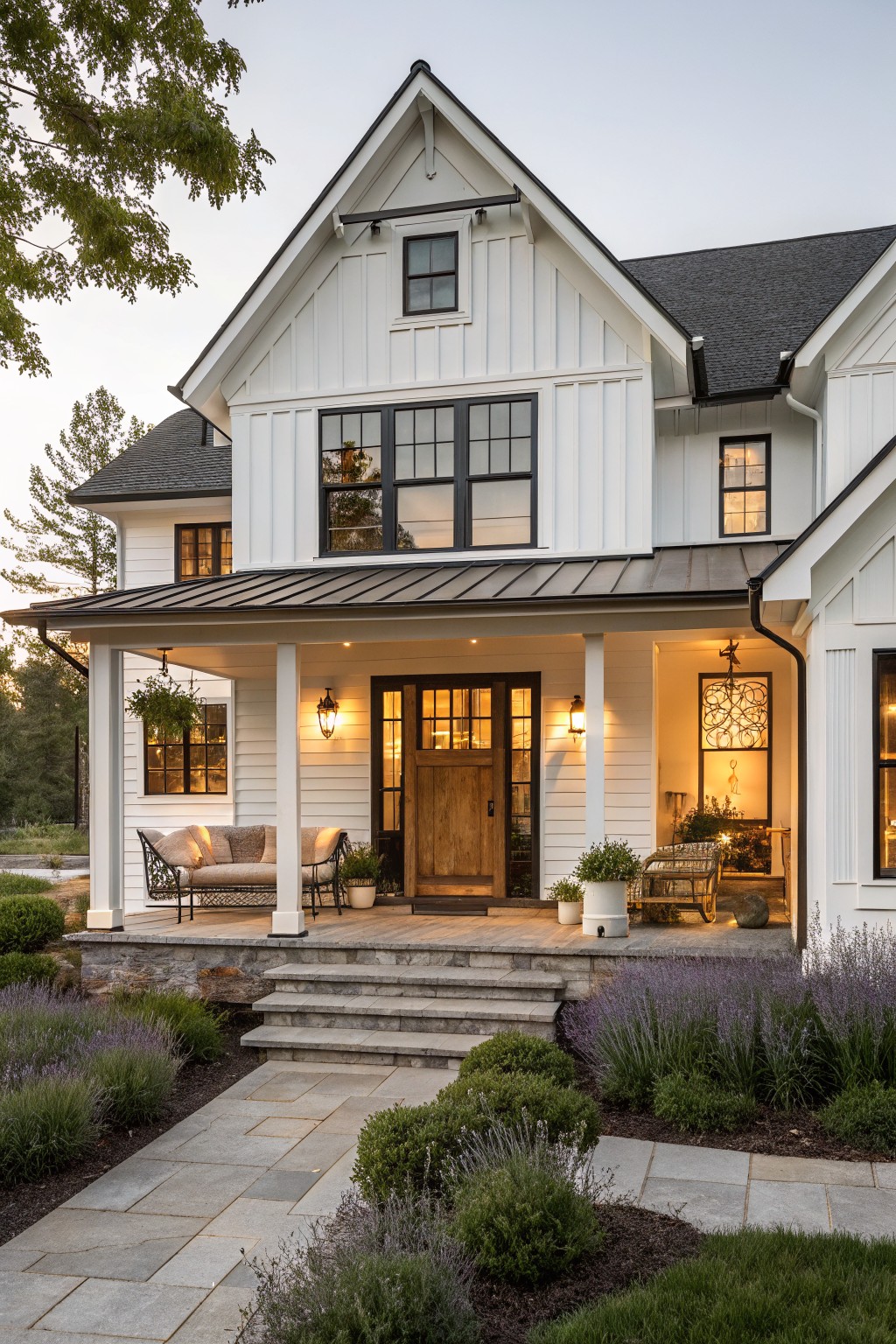 A two-story white board-and-batten sided house with black metal roof, covered front porch featuring wooden double doors, lanterns, wicker seating, stone steps, and lavender plants along a paved pathway.