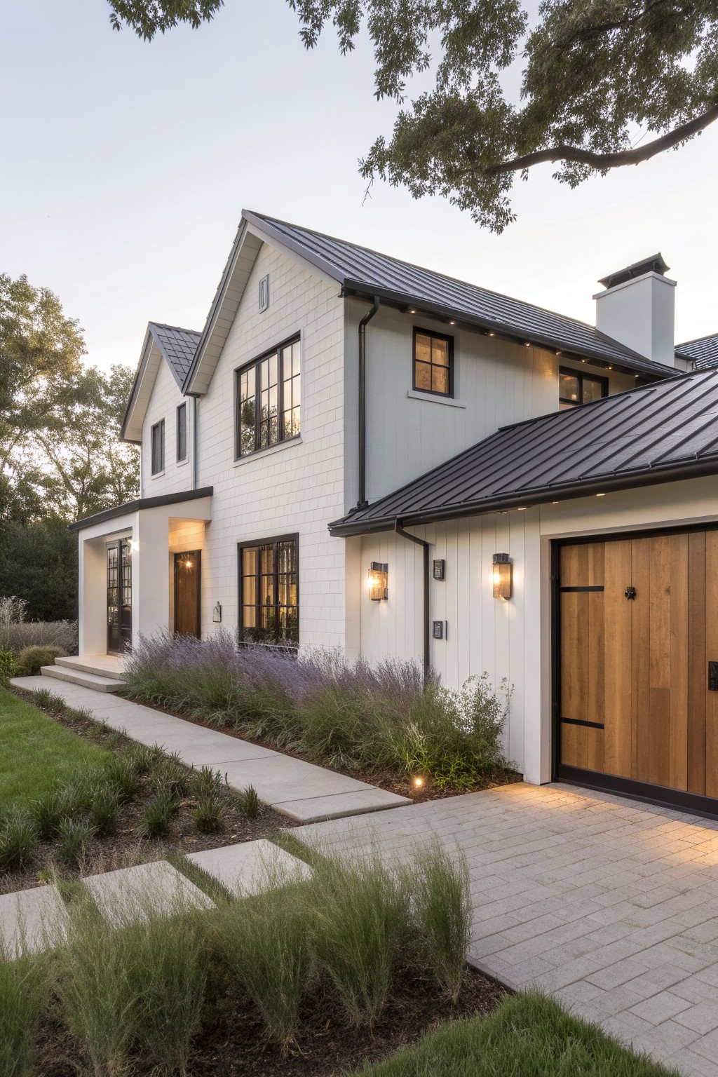 A two-story white house with dark metal roof, wooden garage door, entry porch, and landscaped path with grasses and lavender plants.