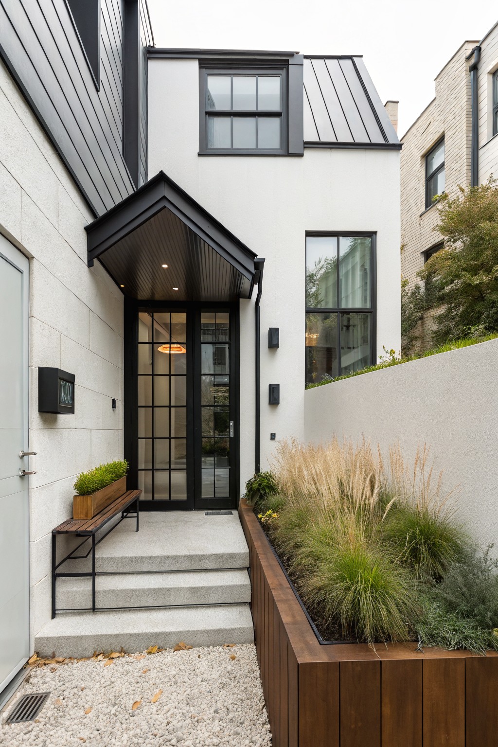White stucco house exterior with black metal framed double glass entry doors under a cantilevered black porch canopy, metal roof, wood bench, planter boxes with grasses, and gravel ground cover.