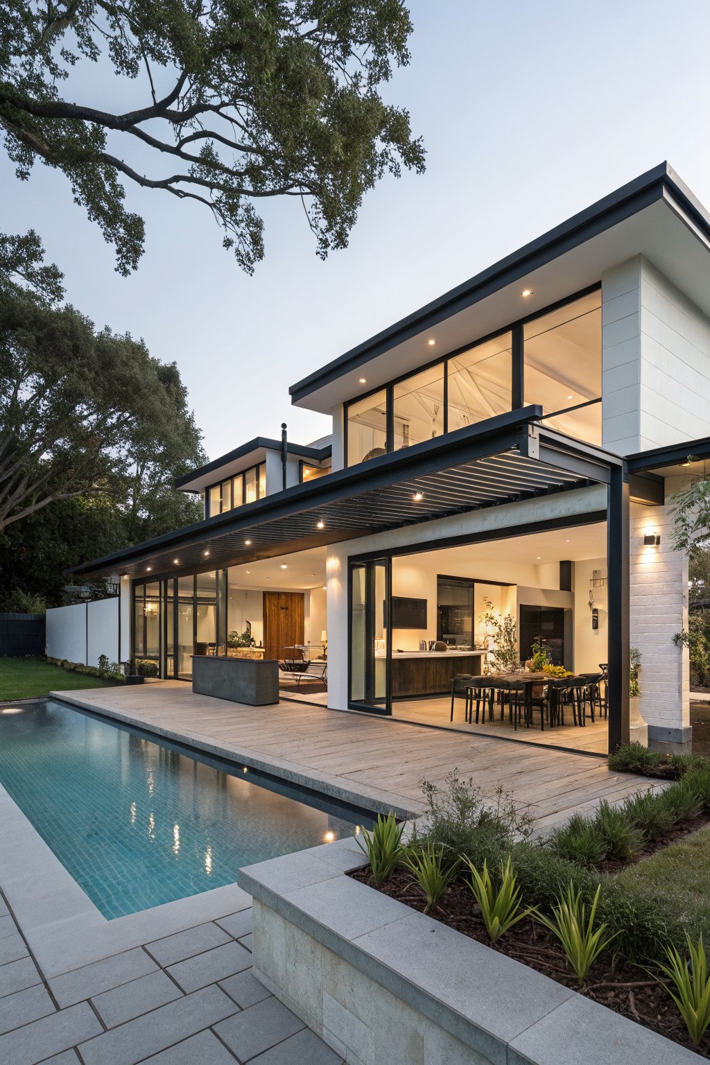 Modern two-story white house exterior with black metal roof and accents, featuring large sliding glass doors opening to a wooden deck, turquoise pool, and surrounding landscaping at dusk.