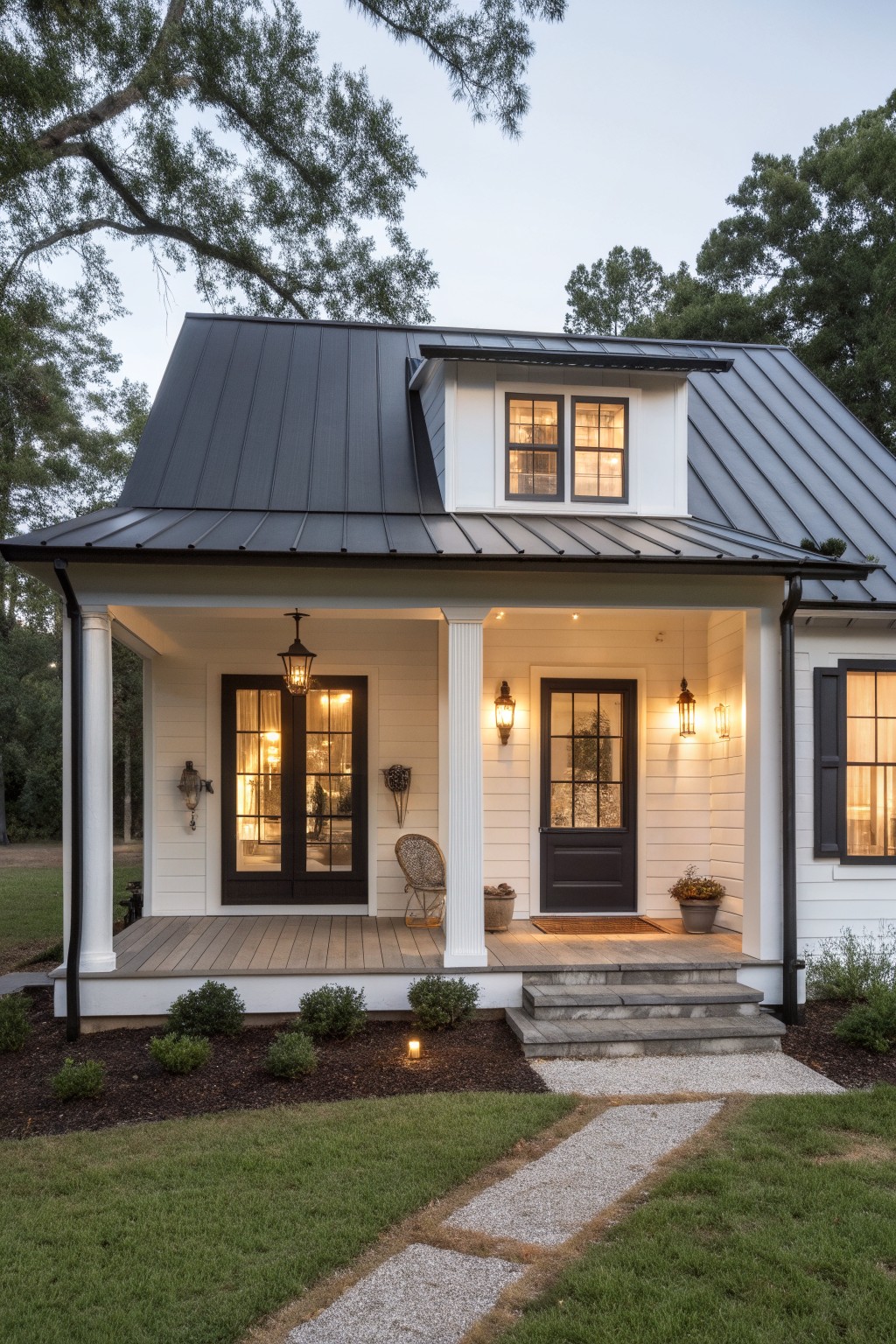 White farmhouse-style house with black metal roof, wraparound porch supported by columns, dark-framed windows and doors, lantern lighting, gravel path, shrubs, and lawn at dusk.