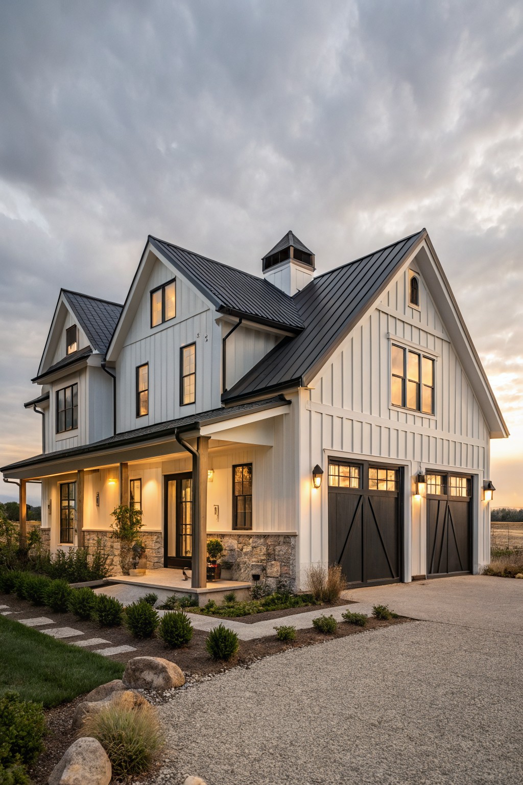 Two-story white board-and-batten house with dark metal gable roofs, black-framed windows and garage doors, covered porch, stone accents, and gravel driveway with landscaping at dusk.