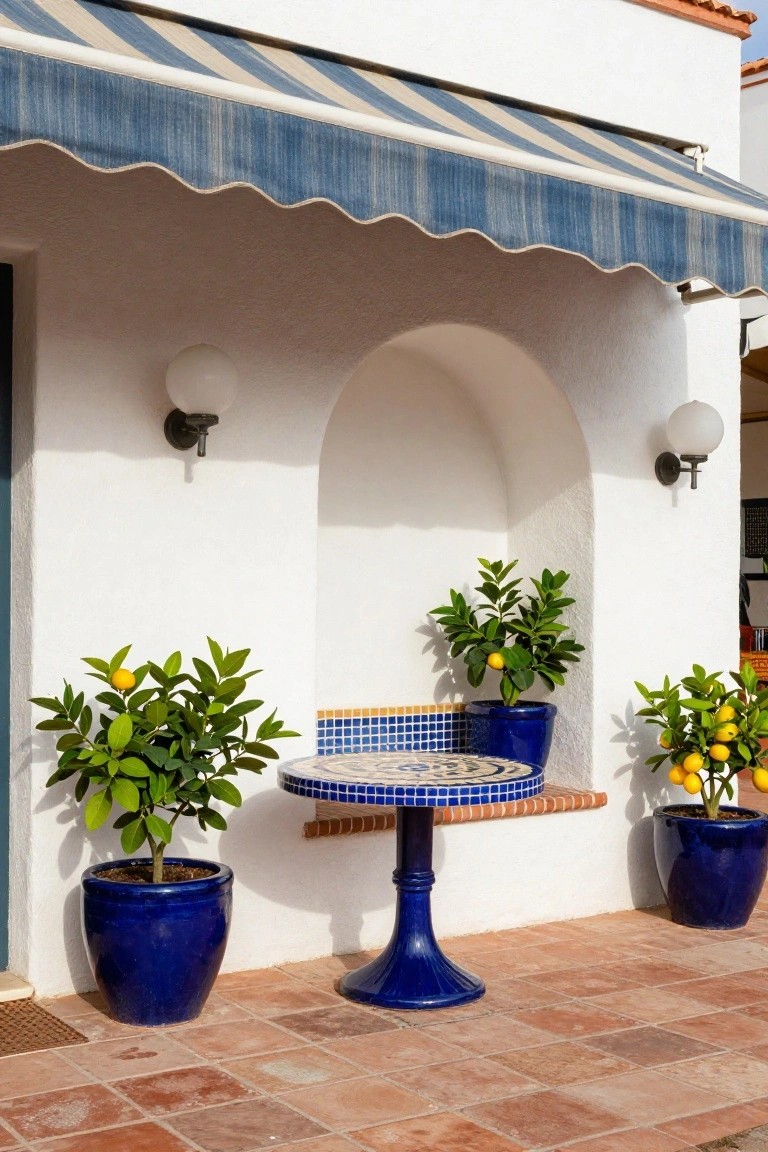 White stucco wall with arched niche holding a blue tiled pedestal table, flanked by blue ceramic pots of lemon trees, under blue and white striped awning on terracotta tile patio.