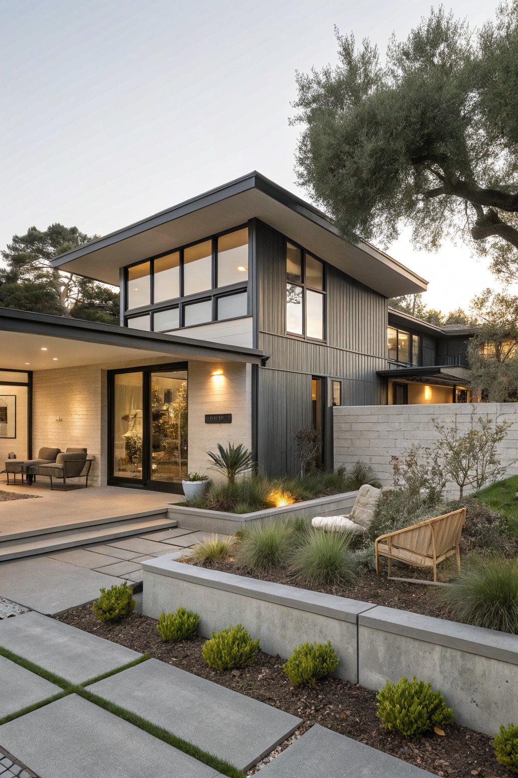 Modern two-story house exterior featuring dark gray vertical siding, large glass windows, beige stone entry with sliding doors, concrete steps and planters, and low landscaping with grasses and shrubs at dusk.