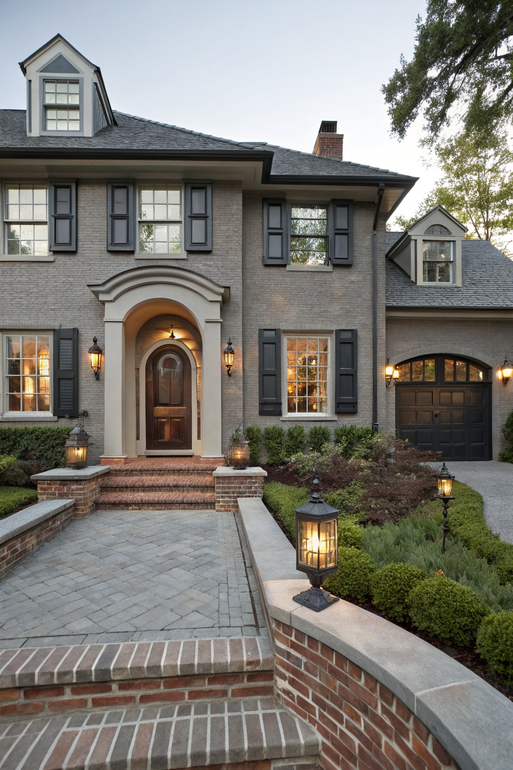 Front exterior of a two-story gray brick house with black shutters, arched entry door, dark garage door, brick steps, and low landscaping with lanterns.