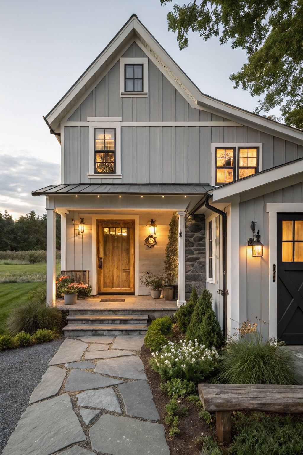 Gray board-and-batten sided two-story house with white trim, covered front porch, wooden door, black garage door, stone accents, lanterns, steps, stone path, and landscaping at evening light.