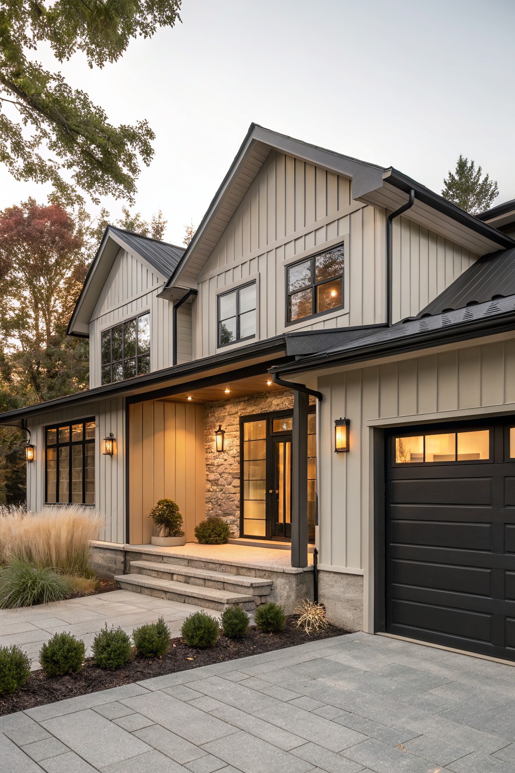 Two-story house exterior with light gray board-and-batten siding, black metal roof and trim, glass front door with stone surround, black garage door, and low landscaping along a stone paver driveway.