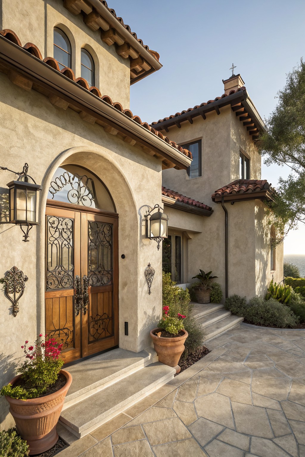 Light beige-gray stucco house exterior featuring an arched wooden double door with wrought iron grilles, flanked by lanterns and potted plants, terracotta tile roof, stone steps, and surrounding landscaping.