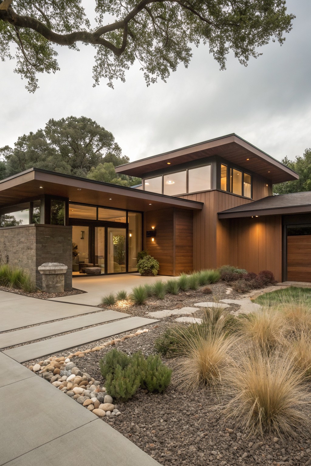 Modern two-story house exterior with brown wood siding, cantilevered upper level, large glass entry doors flanked by stone wall, and gravel landscaping with grasses and stepping stones.