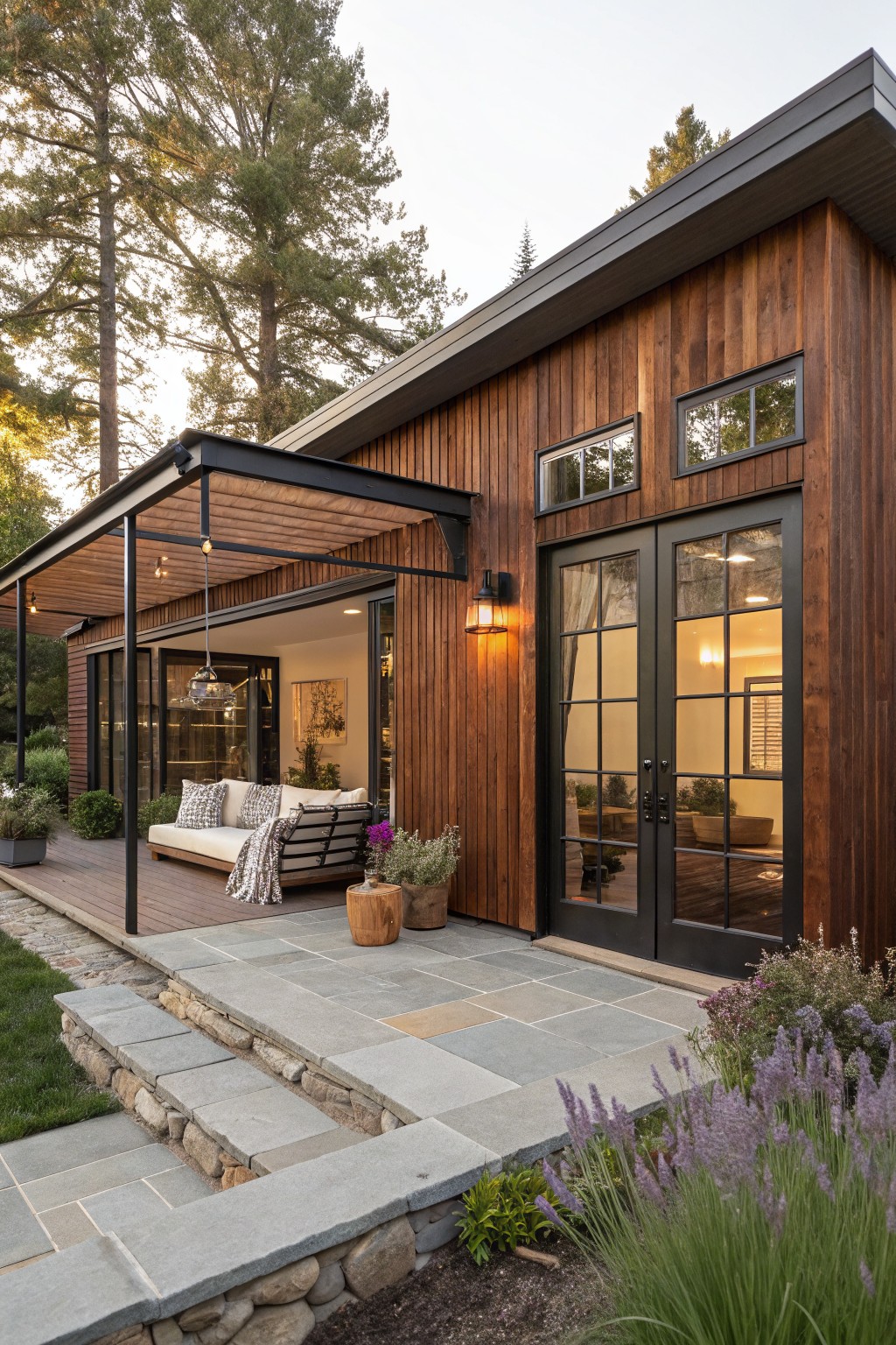 Modern house exterior with vertical brown wood cladding, black-framed glass doors and windows, steel pergola-covered deck with white daybed and plants, stone steps, and surrounding pine trees and lavender.