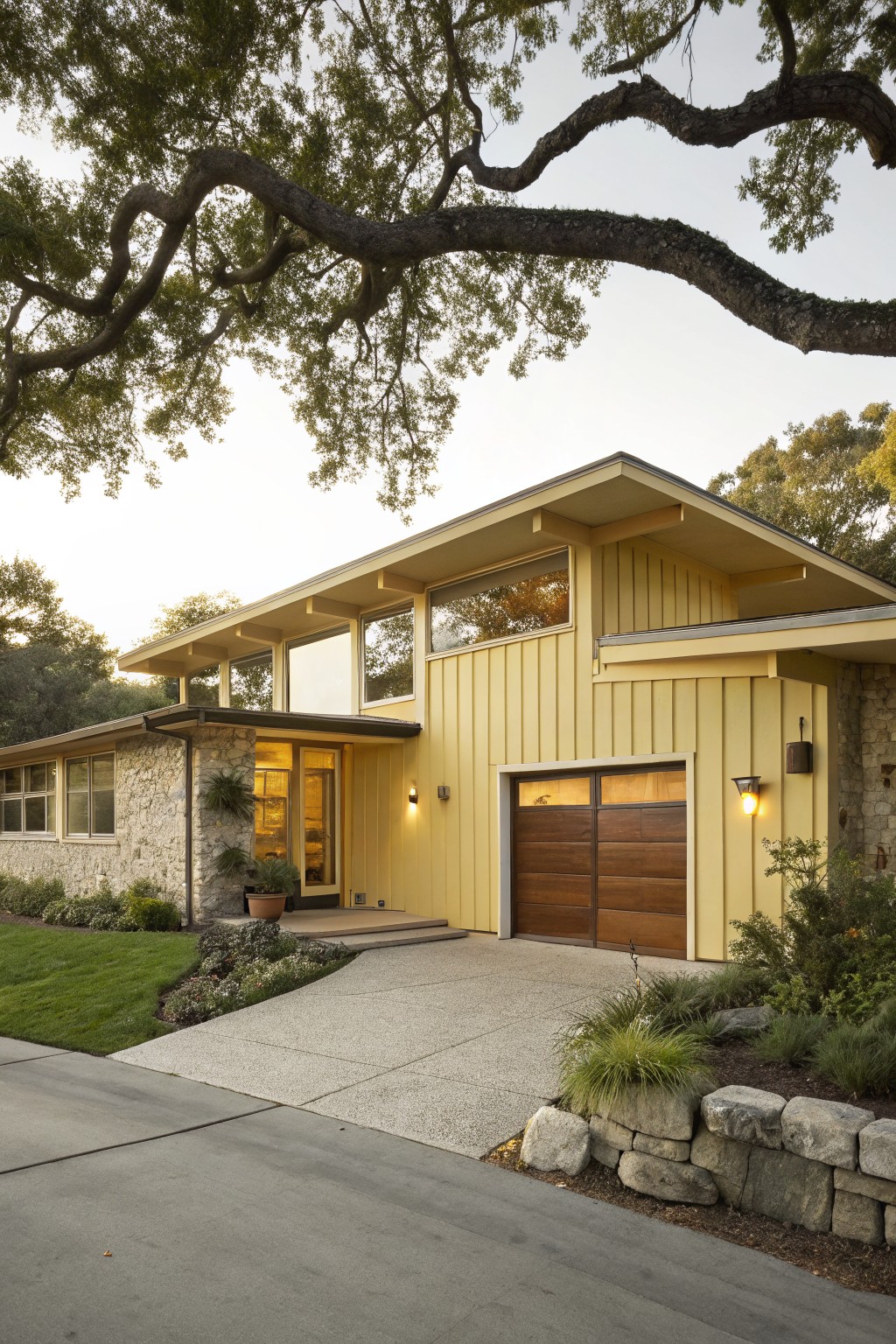 A two-story mid-century modern house with pale yellow vertical siding, sloped roof, large windows, wood garage door, stone base, potted plants at entry, and landscaping including grasses and rocks under oak trees at dusk.