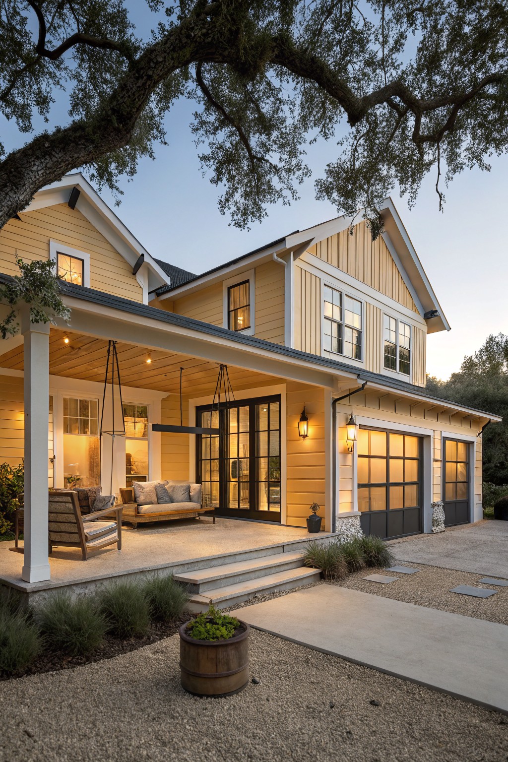 A two-story yellow board-and-batten house with white trim, covered front porch, swing chairs, glass garage doors, lanterns, and gravel landscaping under a large oak tree at dusk.