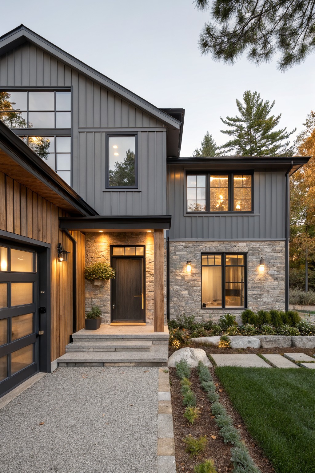 Two-story house exterior featuring warm gray vertical siding, wood-plank garage door, stone entryway with dark wood door, large windows, and gravel path with landscaping at dusk.