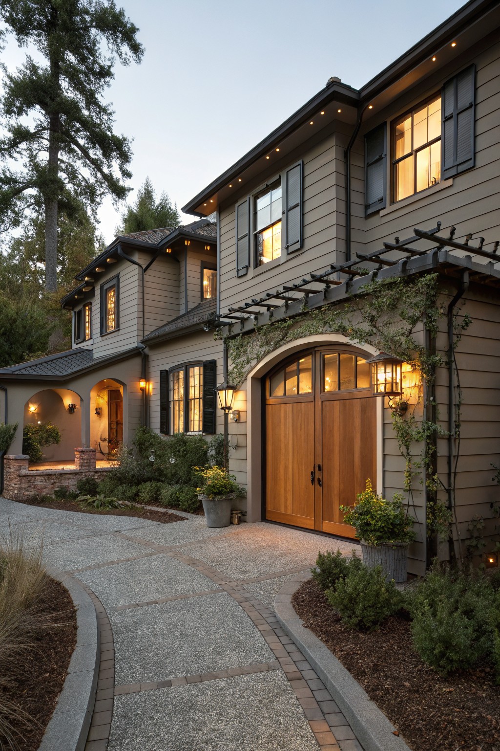 Two-story house exterior with warm gray horizontal siding, large wooden double garage doors, arched windows, lit lanterns, vine-covered trellis, and gravel pathway bordered by plants at dusk.