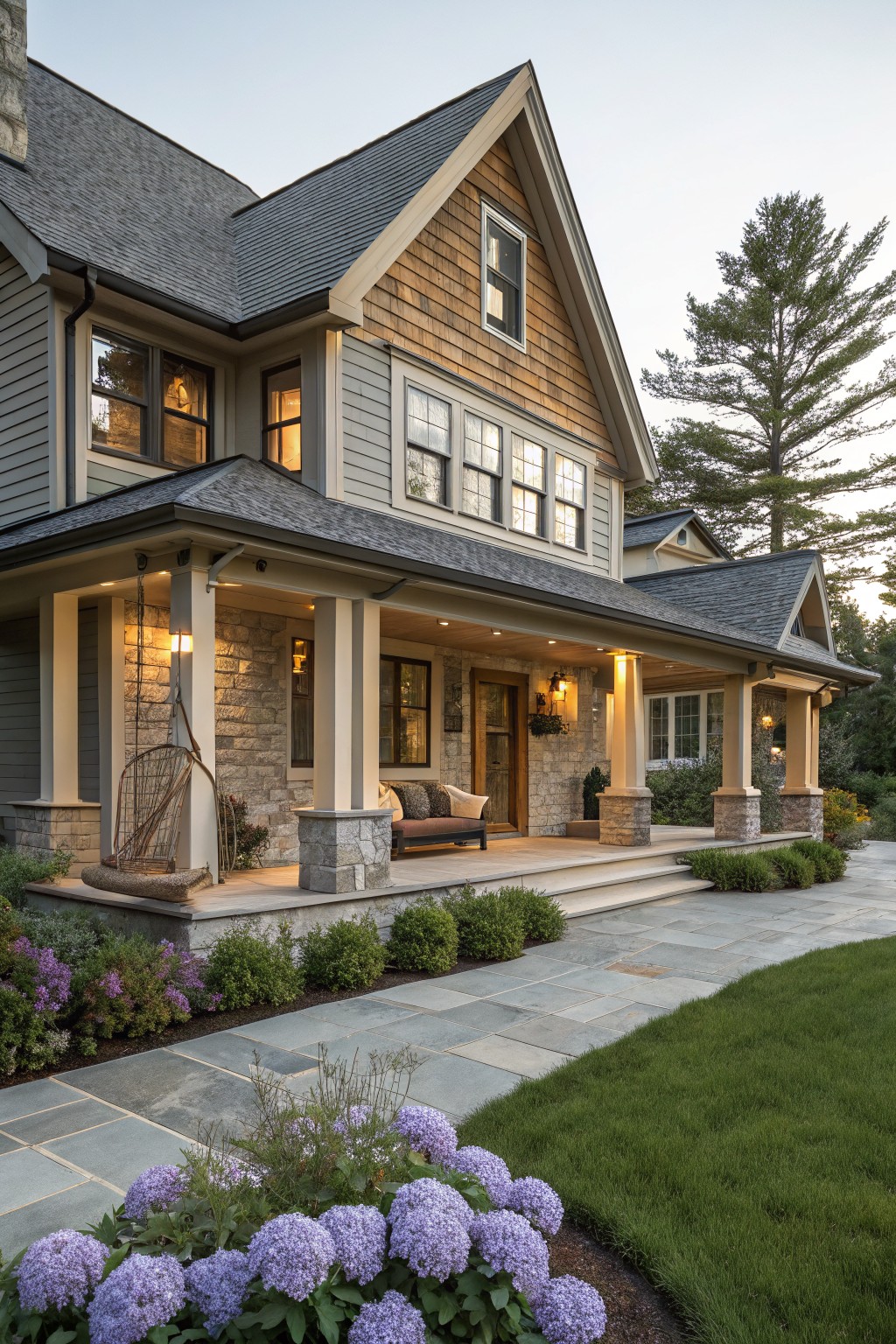 Two-story house with warm gray siding, wood shingle gables and porch roof, stone porch foundation and columns, hanging swing and bench on porch, bluestone walkway edged by shrubs and hydrangeas, tall pine trees nearby at dusk.