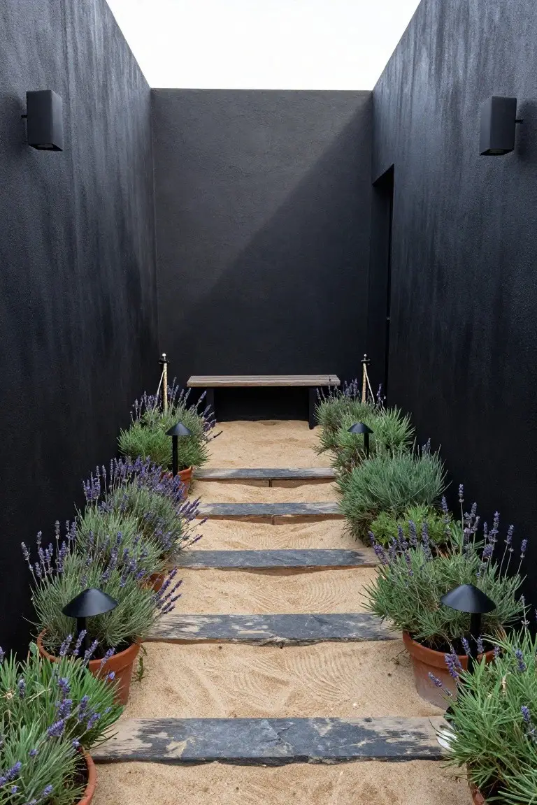 Narrow outdoor path between tall matte black stucco walls with sand ground cover and irregular dark stone slab steps leading to a black door, lined with lavender plants in terracotta pots and black mushroom lights, wooden bench midway.