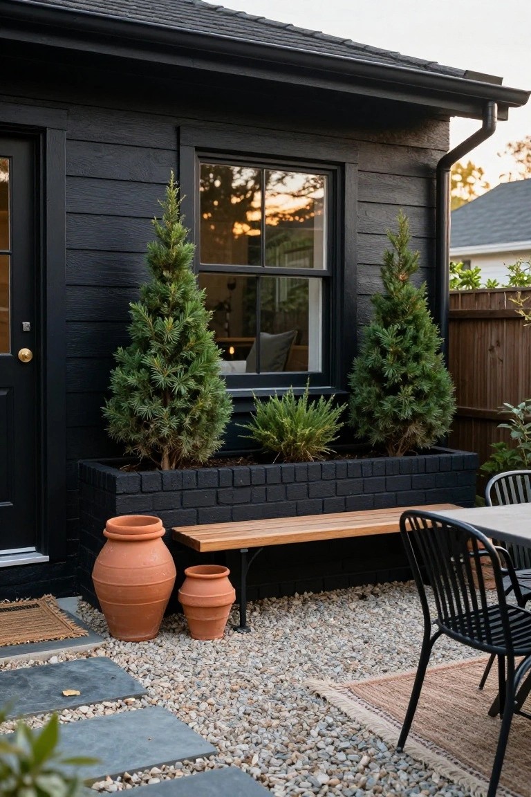 Black wood-clad exterior wall of a shed-like building with a black door and window, adjacent black brick raised planter box holding two small evergreen trees and a central shrub, large terracotta pots nearby, wooden bench, gravel ground cover, slate steps, and black metal patio chairs at a table.
