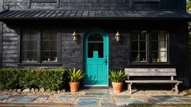 Two-story black clapboard house exterior featuring a turquoise paneled front door with brass hardware, flanked by lantern lights, potted ferns, a wooden bench, and a stone path on gravel landscaping.