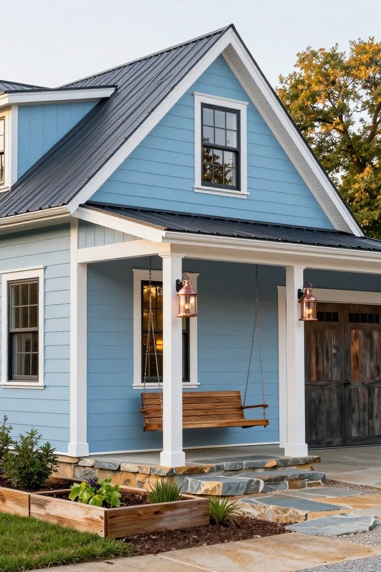Light blue gable-front house with black metal roof, white trim, covered porch featuring wooden swing and lanterns, wooden garage doors, stone entry steps, and raised garden beds.