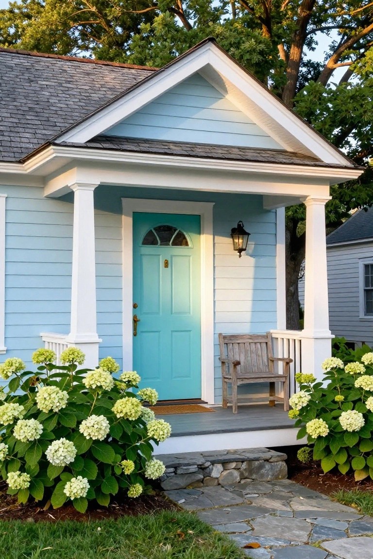 Light blue clapboard house with white trim and porch columns, turquoise arched front door, lantern light, wooden bench, large white hydrangea bushes, and stone steps leading to the entry.