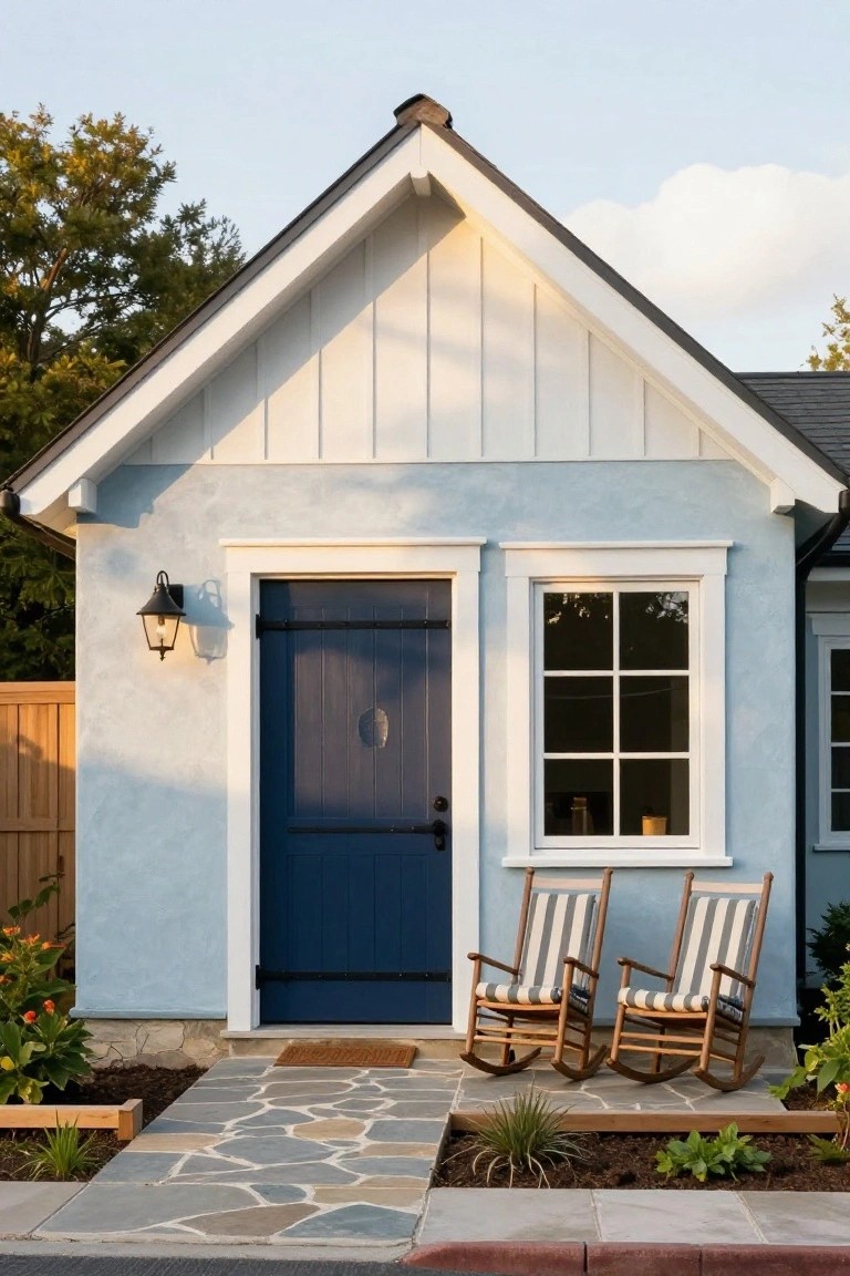 Light blue stucco cottage house exterior with gabled roof, white board-and-batten siding, navy paneled door with lantern light, six-pane window, two striped wooden rocker chairs on porch, stone walkway, plants in beds, wooden fence, and asphalt street.