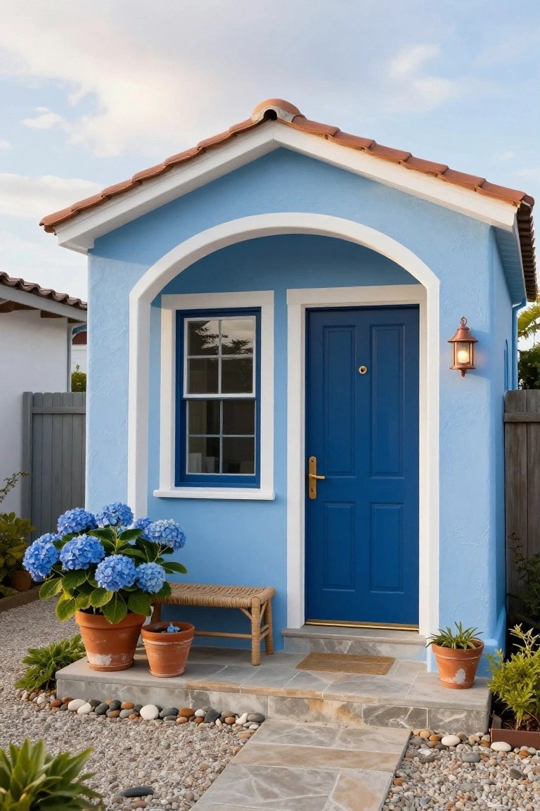 Small light blue stucco house with white-trimmed arched entryway, dark blue front door, blue hydrangeas in terracotta pots, wooden bench, lantern light, pebble ground cover, and terracotta tile roof.