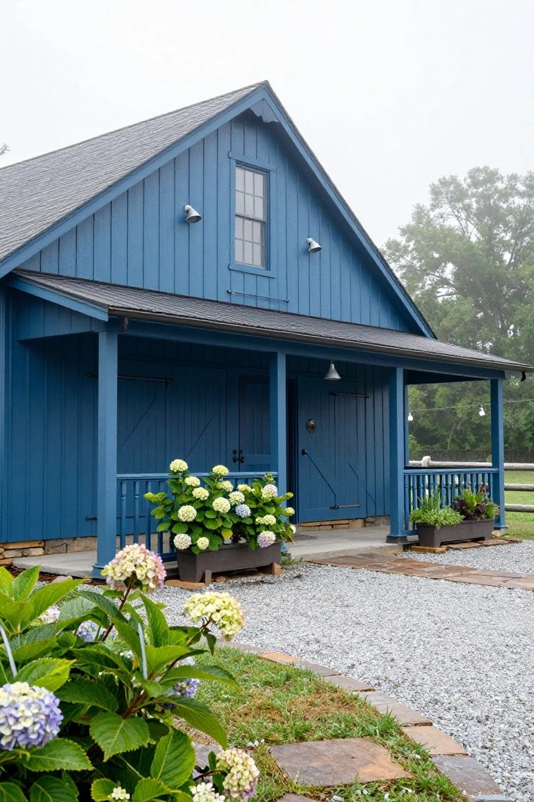 Navy blue board-and-batten barn-style building with gabled roof, double garage doors, covered porch with railings, white lanterns, potted white hydrangeas, gravel driveway, stone paths, and surrounding greenery on a misty day.