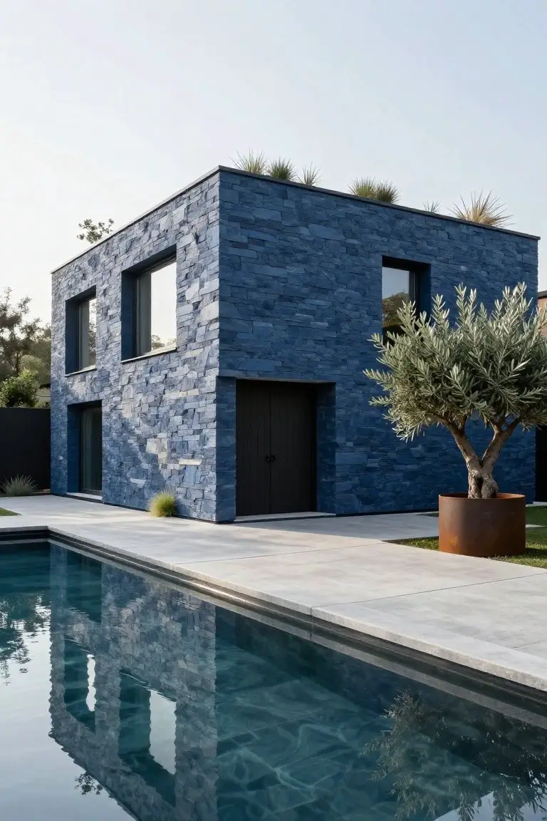 A modern corner house with blue stone cladding, narrow black-framed windows, dark garage door, olive tree in large rust-colored pot, and adjacent pool reflecting the facade on a paved terrace.