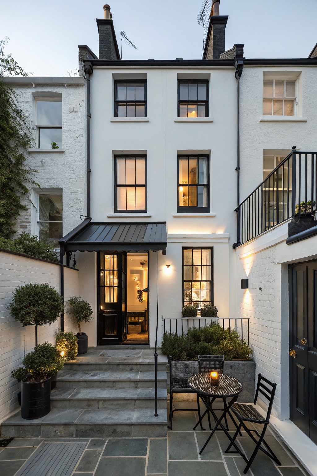 White terraced houses viewed from a small front courtyard at dusk, with the central house showing black-framed windows, black metal awning over glass entry doors, stone steps, potted plants, and a bistro table with chairs.