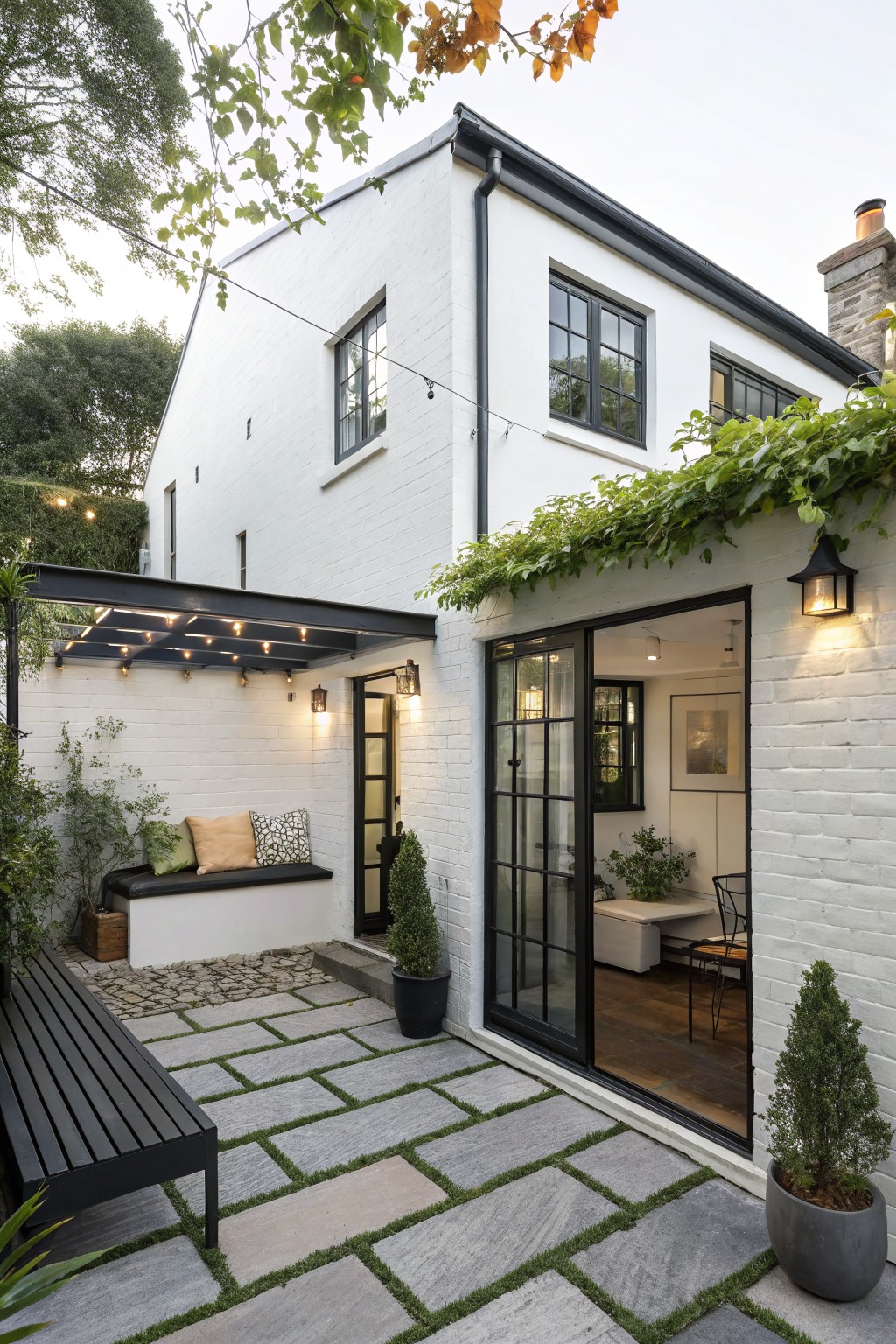 White brick house exterior with black-trimmed windows and sliding glass doors, black metal pergola with string lights over a stone-paved patio featuring a built-in bench, potted plants, and boxwood shrubs.