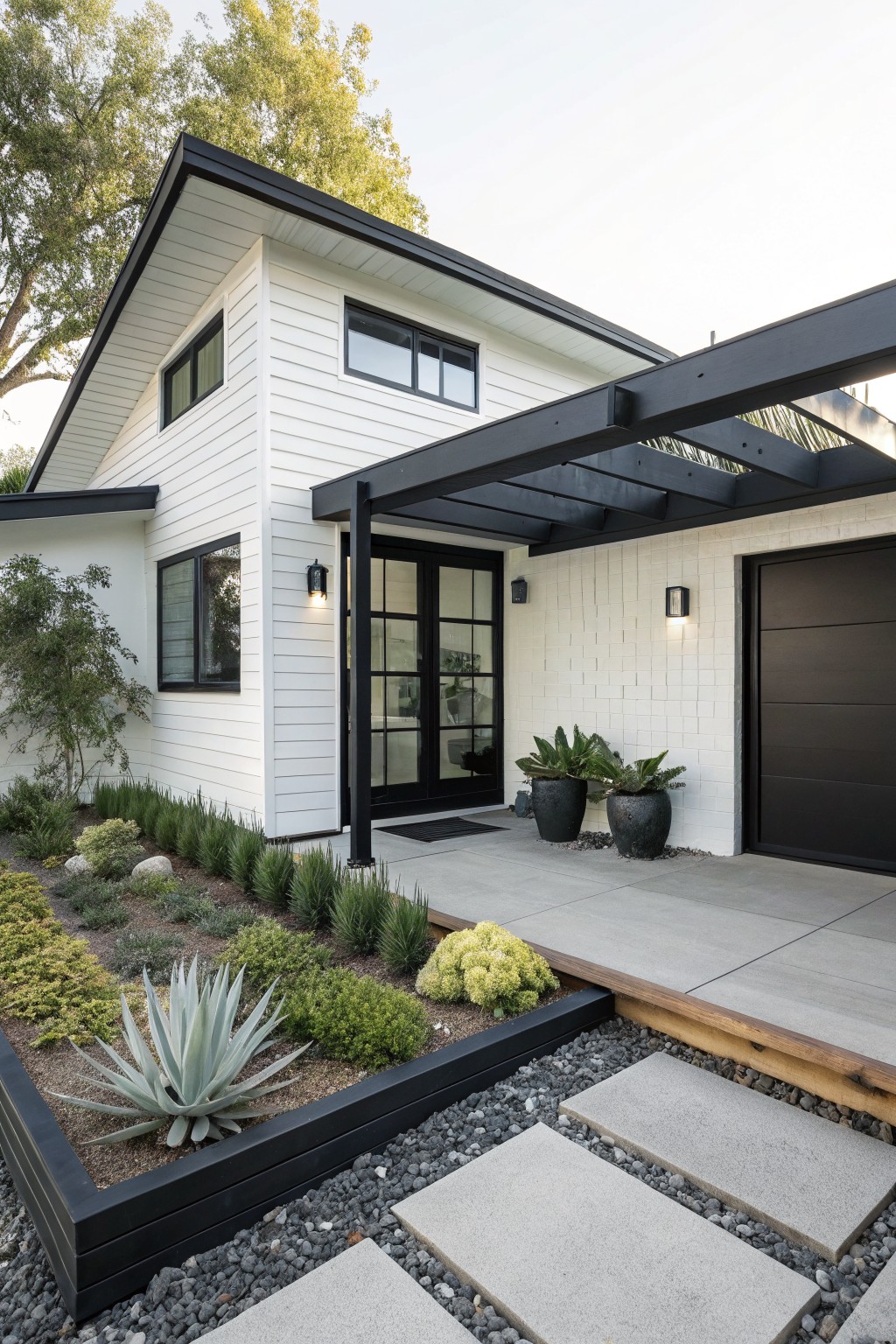 White shiplap house exterior with black metal pergola extending over front entry doors and garage, black-framed windows, potted plants, raised landscaped bed with succulents, and concrete paver path.
