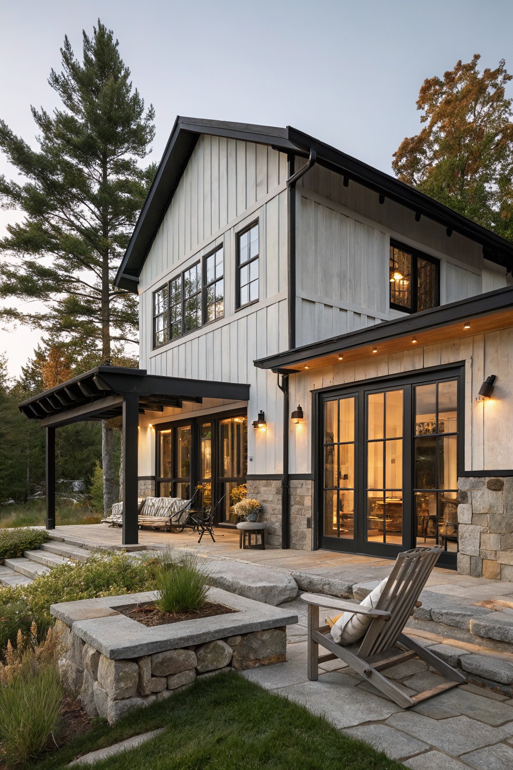 Side view of a two-story house with white board-and-batten siding, black trim on large multipane windows and doors, covered porch with black beams and posts, stone foundation and steps, wooden deck with chairs and plants, surrounded by pine trees and landscaping at dusk.