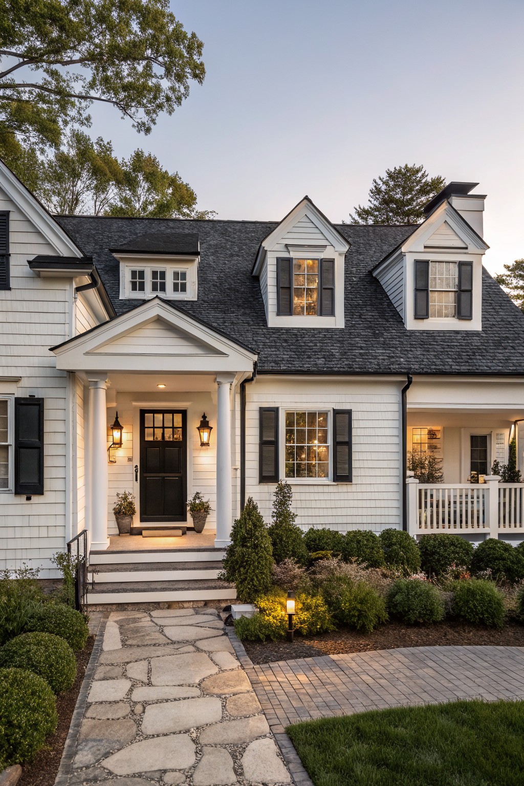 White two-story house with black roof shingles, shutters, front door, and trim on a porch with columns, flanked by landscaping and a stone pathway leading from the street at dusk.