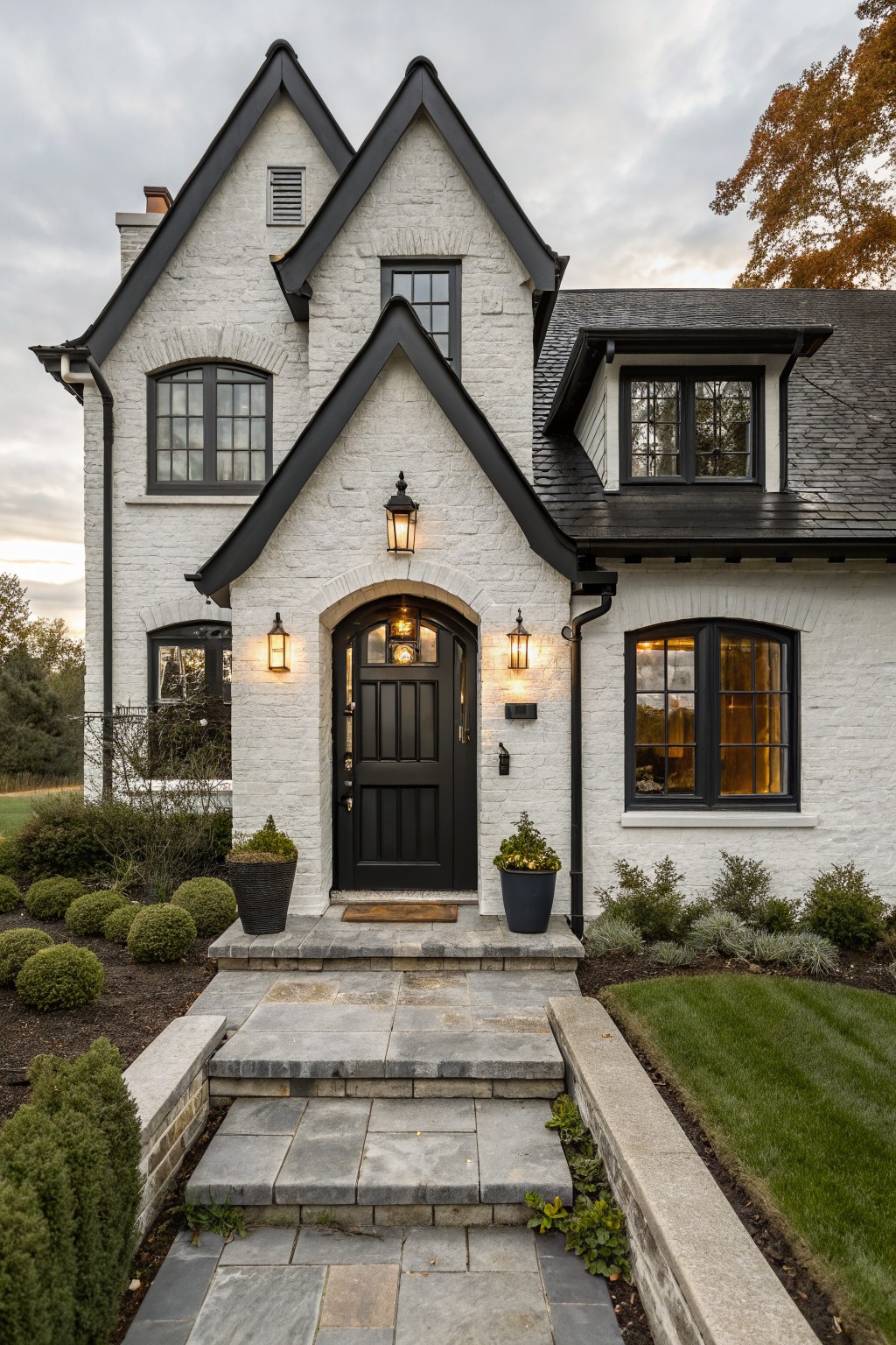 White brick two-story house with black trim on gabled roofs, multi-pane windows, arched entry door flanked by lanterns, stone steps, potted plants, shrubs, and lawn.