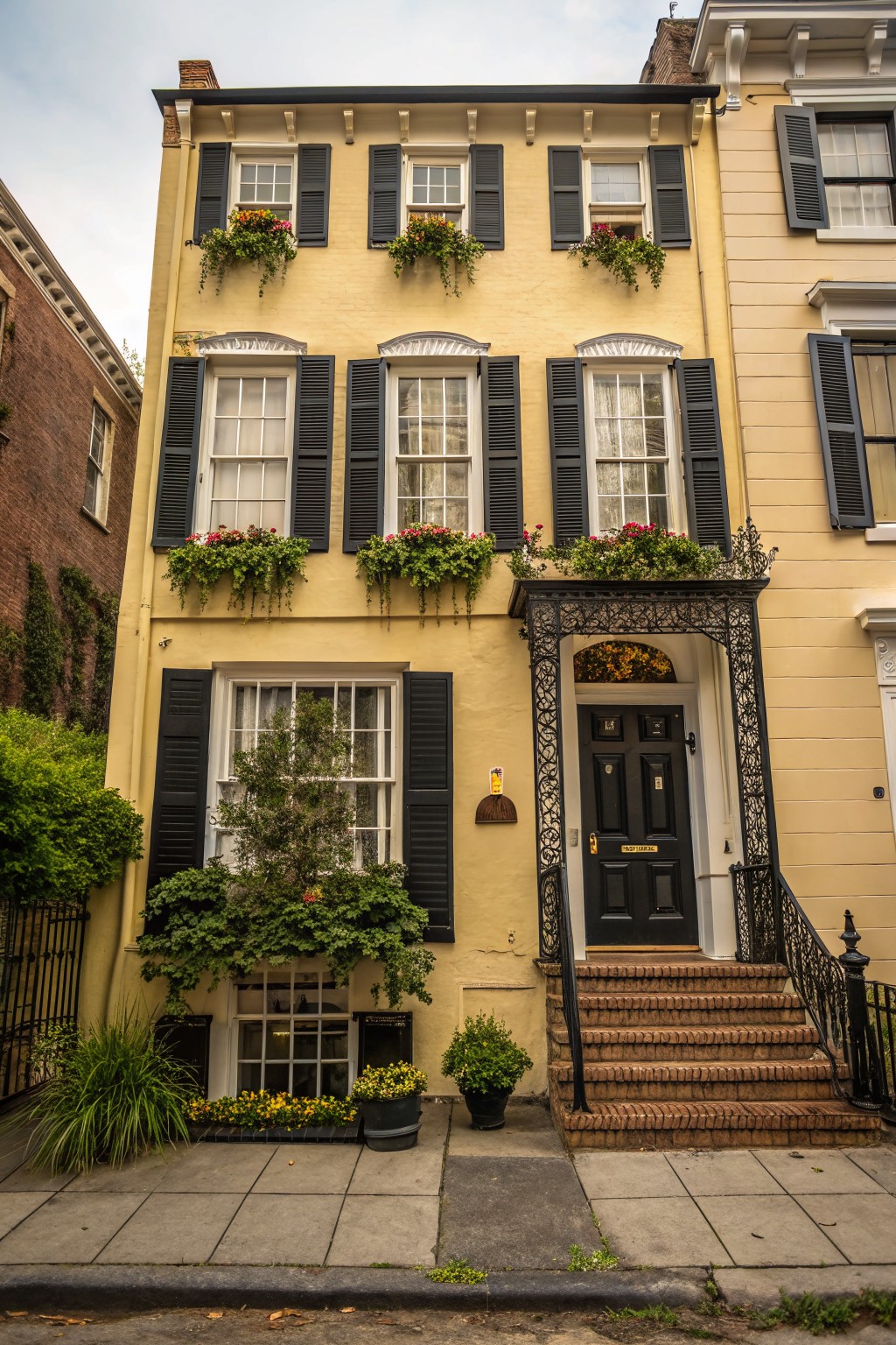 A two-story pale yellow townhouse with black louvered shutters on multi-pane windows, flower boxes overflowing with plants, a black front door in a wrought iron entry archway with brick steps, flanked by greenery and potted plants on a sidewalk.