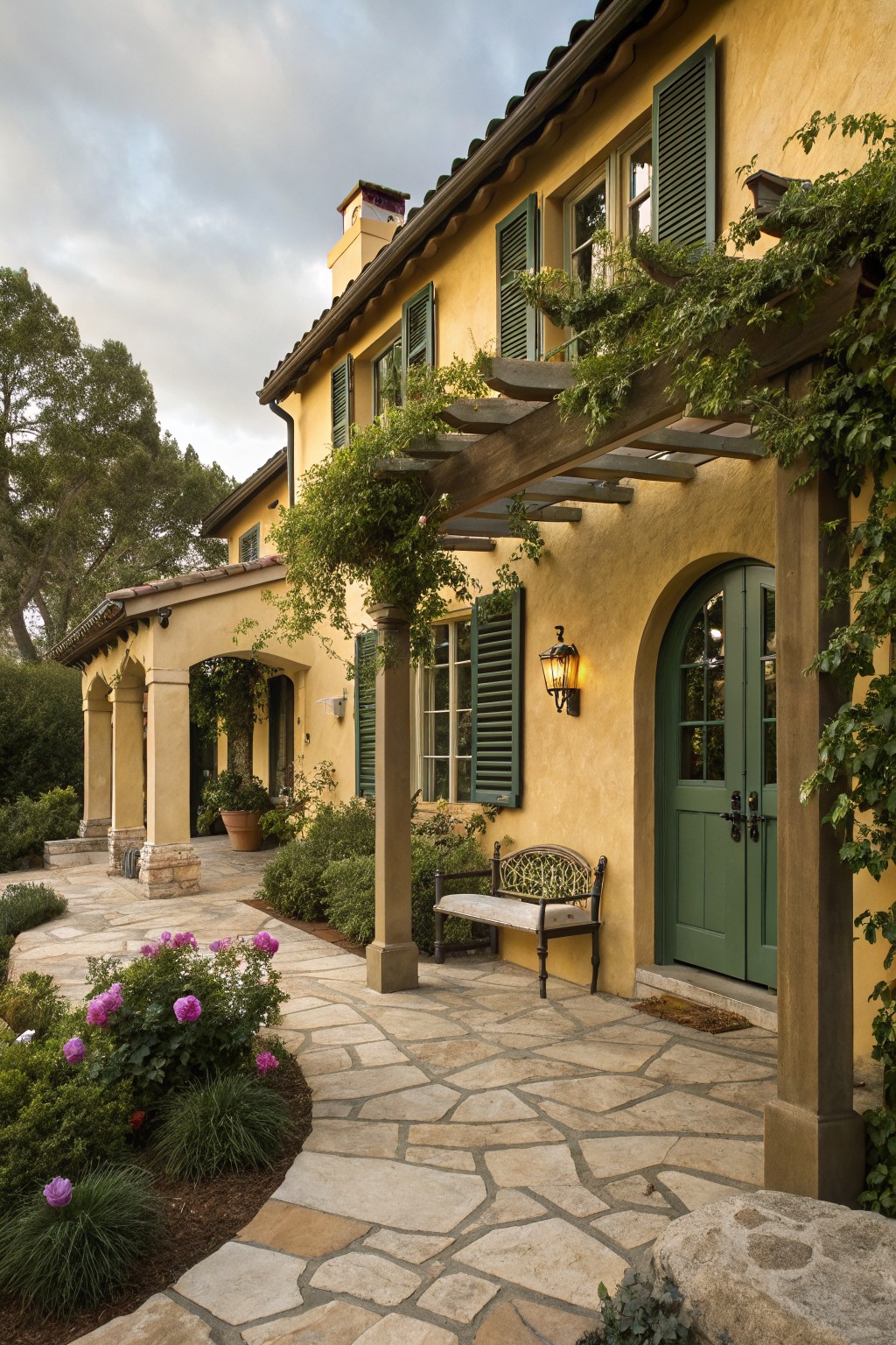 Yellow stucco house exterior featuring green shutters on windows, an arched green entry door, wooden pergola with vines, stone pathway lined with plants and flowers, and a bench under the pergola.