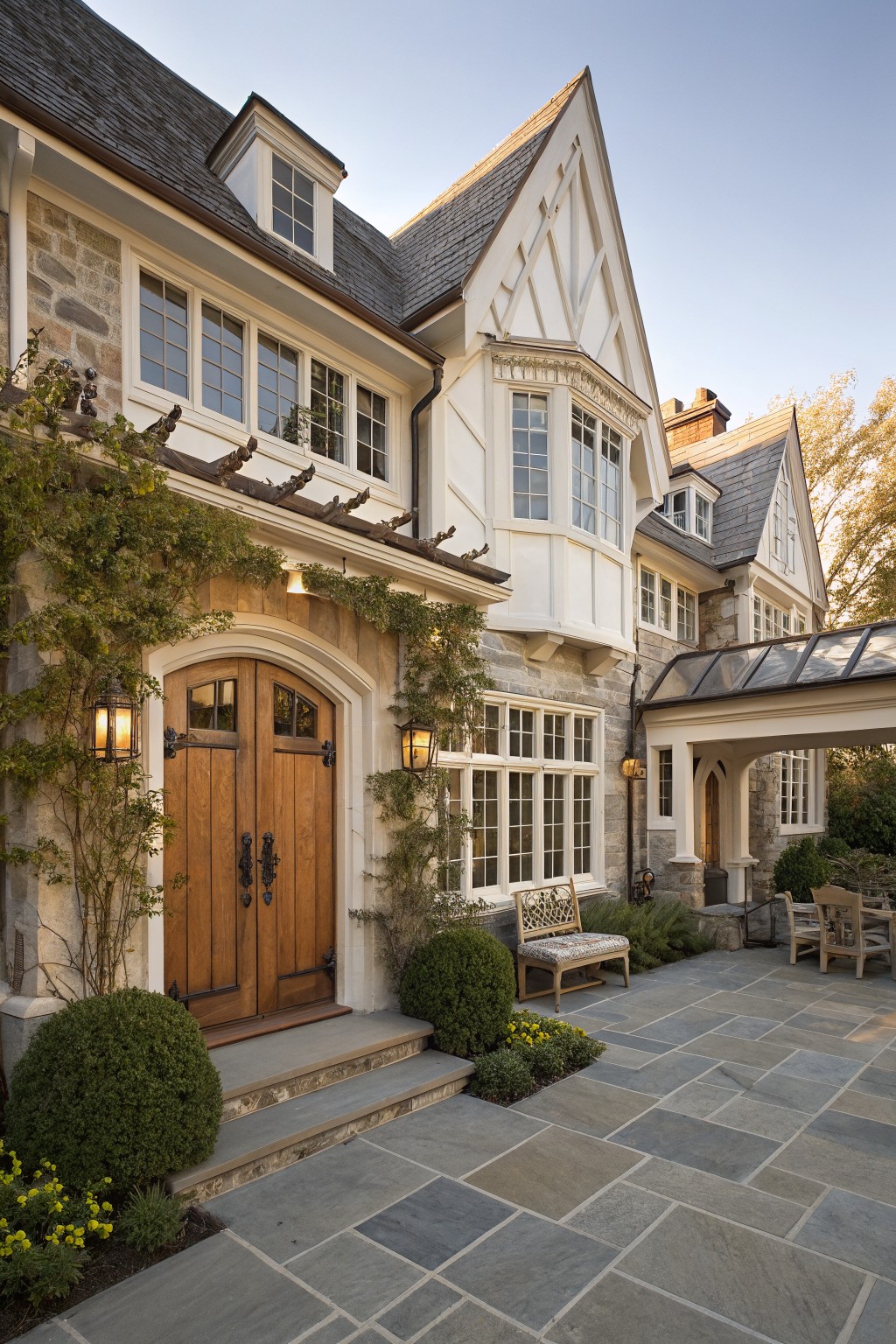 Stone house exterior with tall arched double wooden front doors, flanked by lanterns and climbing vines, white trim on windows and gables, and a stone patio with benches.