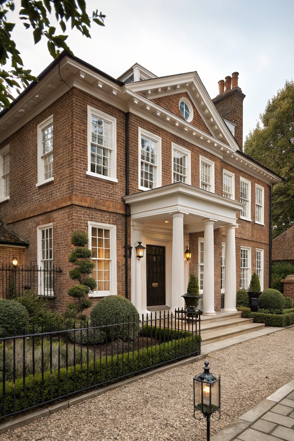 A two-story red brick house exterior with white window trim and a white pedimented portico supported by four columns at the front entrance, flanked by topiary plants, black iron fencing, and a gravel driveway.