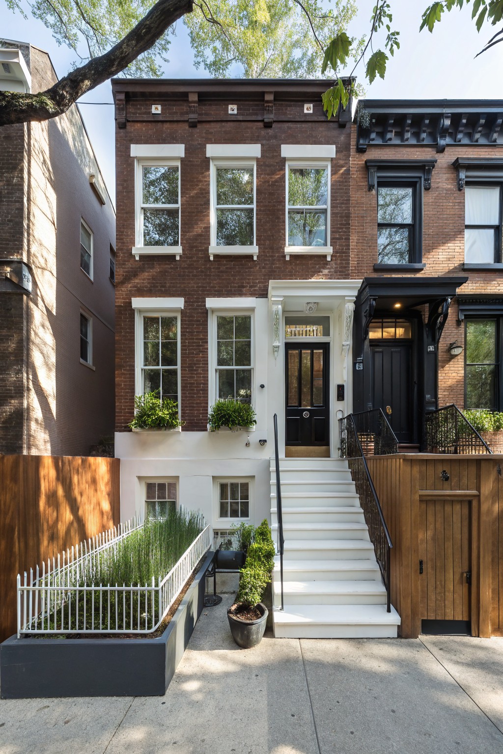 Three attached brown brick townhouses with white window frames and trim, central house featuring a white stoop with black door, potted plants, and front fence with greenery on a sunny street.
