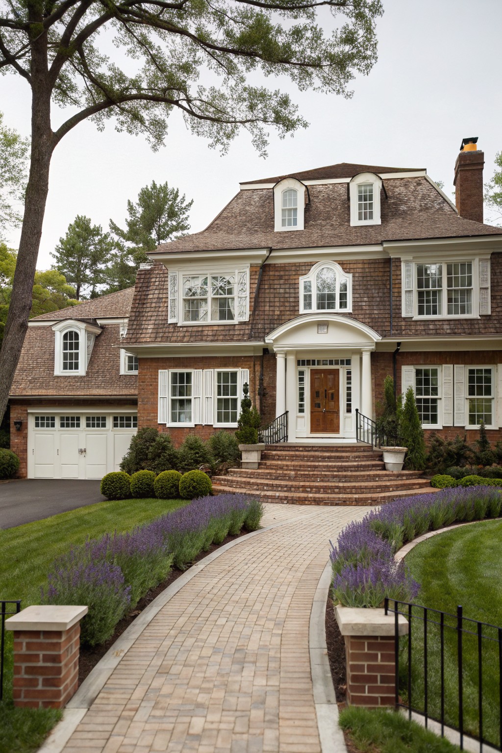 Two-story house exterior with brown brick base, shingled roof and gables, white window trim and columned portico over wood front door, brick steps, and curved brick pathway lined with lavender plants.